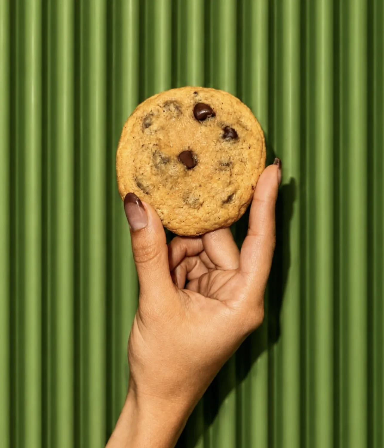 Mano sosteniendo una galleta con chispas de chocolate contra una pared verde acanalada