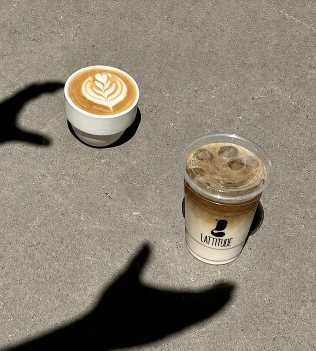 Toma cenital de un latte con arte y un café helado para llevar, con la sombra de una mano sobre el pavimento