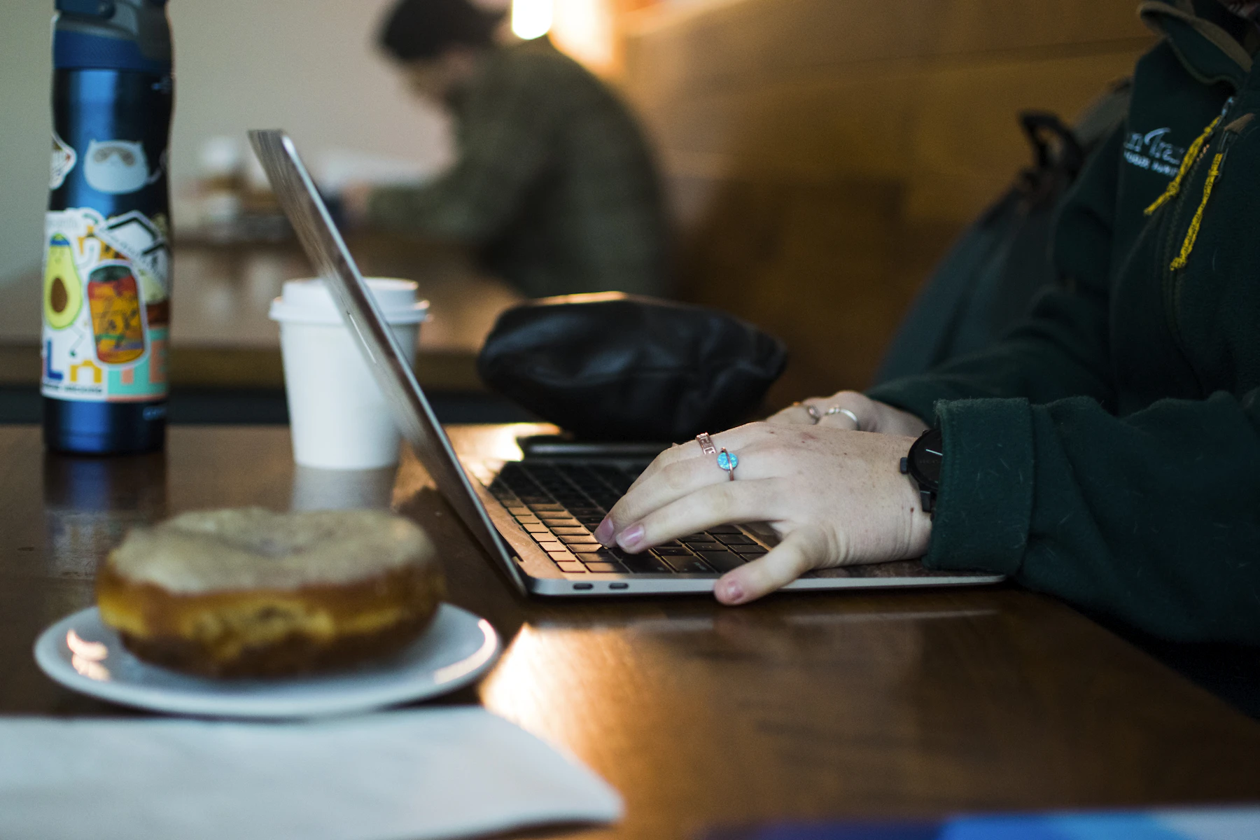 Persona trabajando en una laptop en Lattitude Coffee, con un café, un termo y una rebanada de pastel sobre la mesa
